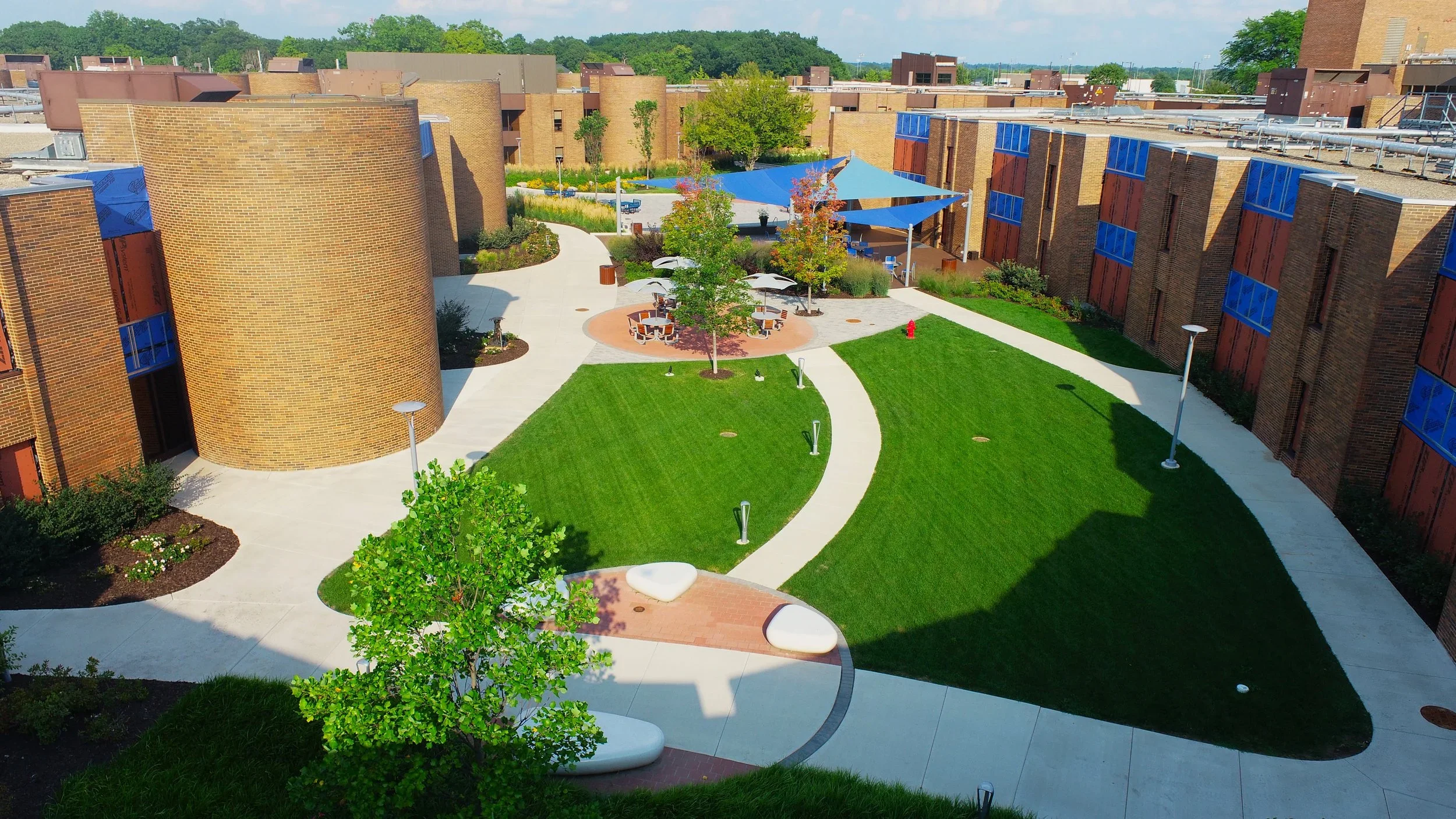A courtyard between brick residential buildings with green lawns, paved walkways, trees, outdoor seating area with umbrellas, and lighting fixtures.