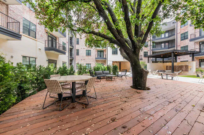Outdoor courtyard with a large tree, patio furniture, and a brick-paved ground surrounded by multi-story apartment buildings.