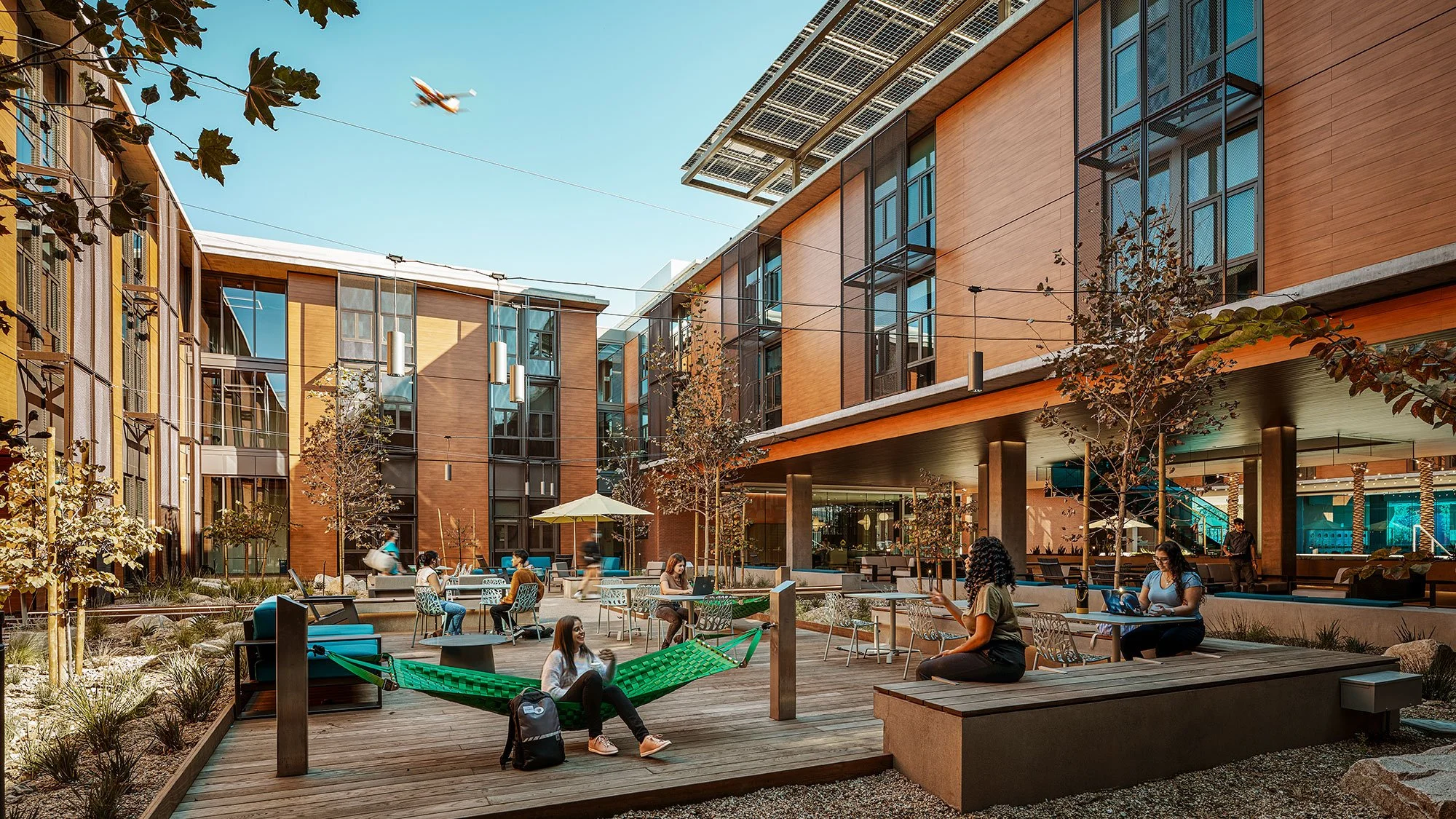 People relaxing and working in an outdoor courtyard with benches, tables, and a hammock, surrounded by modern buildings and trees.