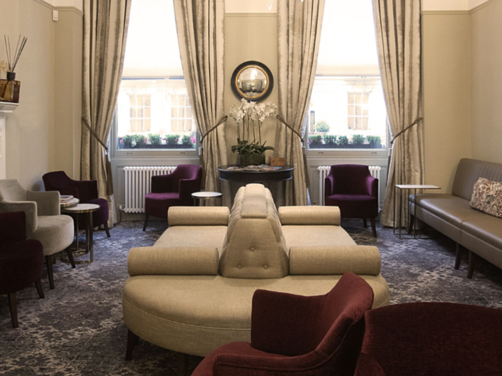 Living room with beige, gray, and burgundy furniture, patterned carpet, large windows with cream curtains, and decorative plants.