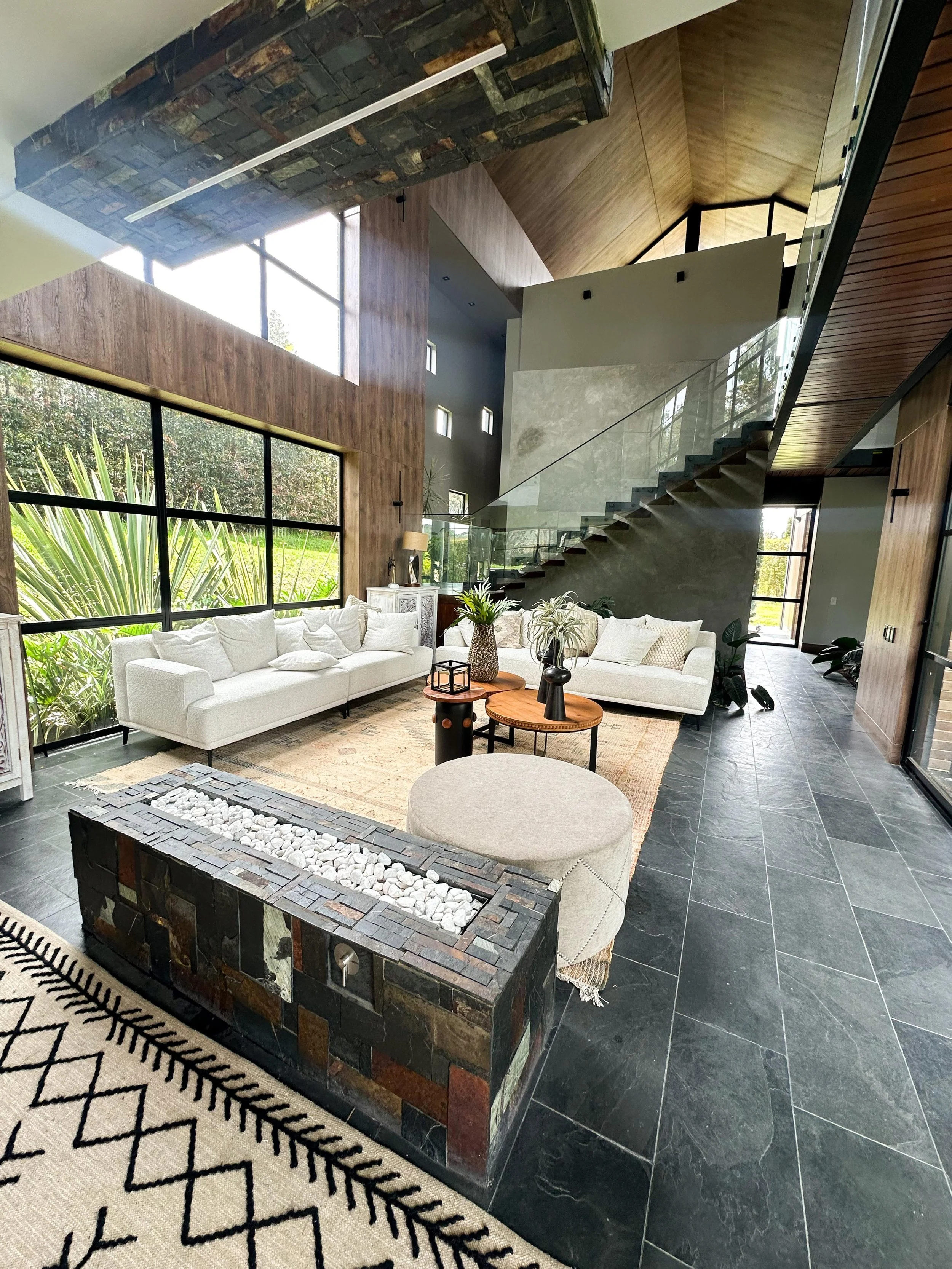 Sala moderna con dos sofas blancos, mesas de madera, plantas, una pared de piedra y ventanas grandes con vista a un jardín