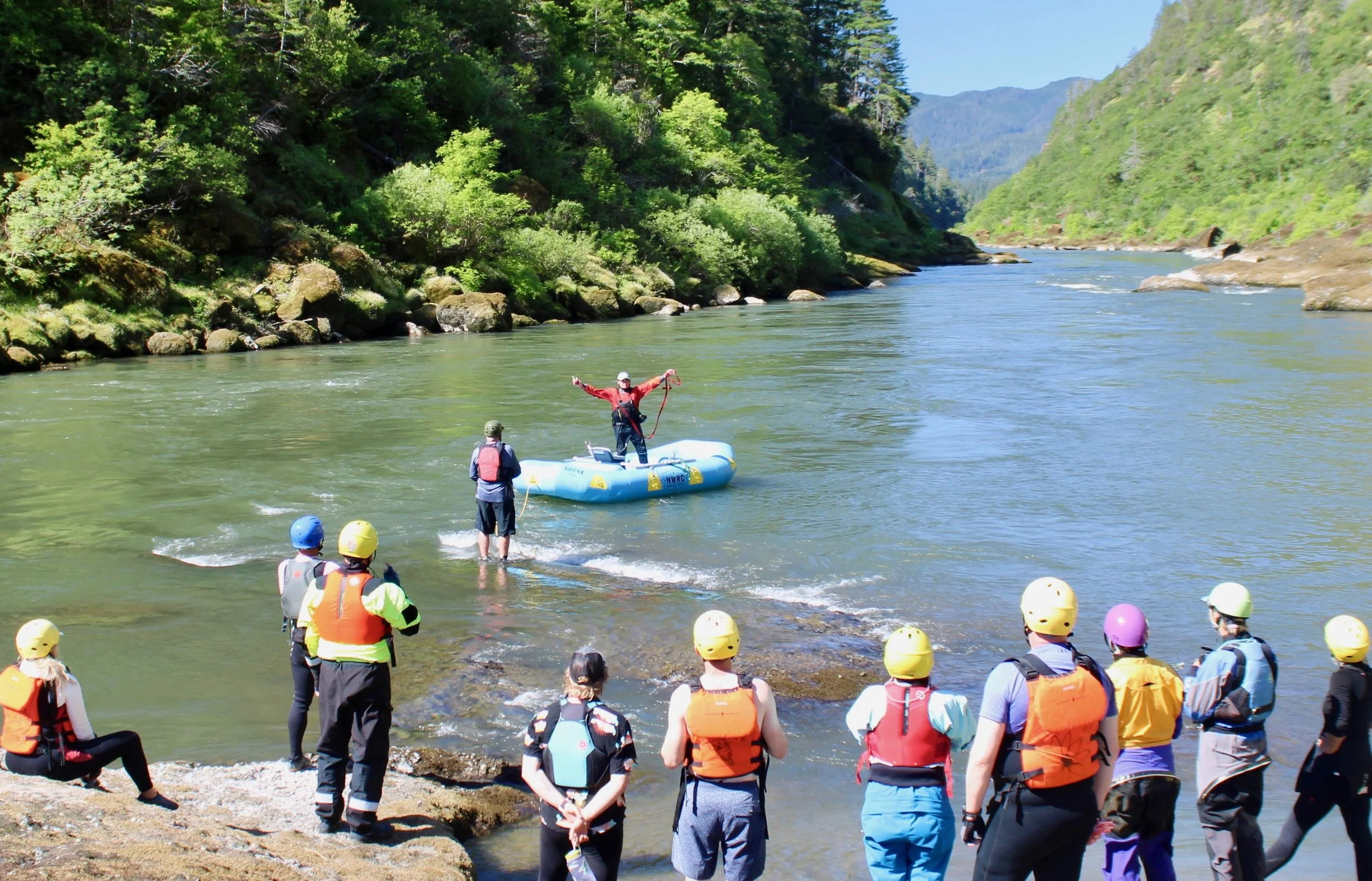 People in life jackets and helmets watching a person in a red jacket holding a rope and standing on an inflatable raft in a river surrounded by green hills and trees.