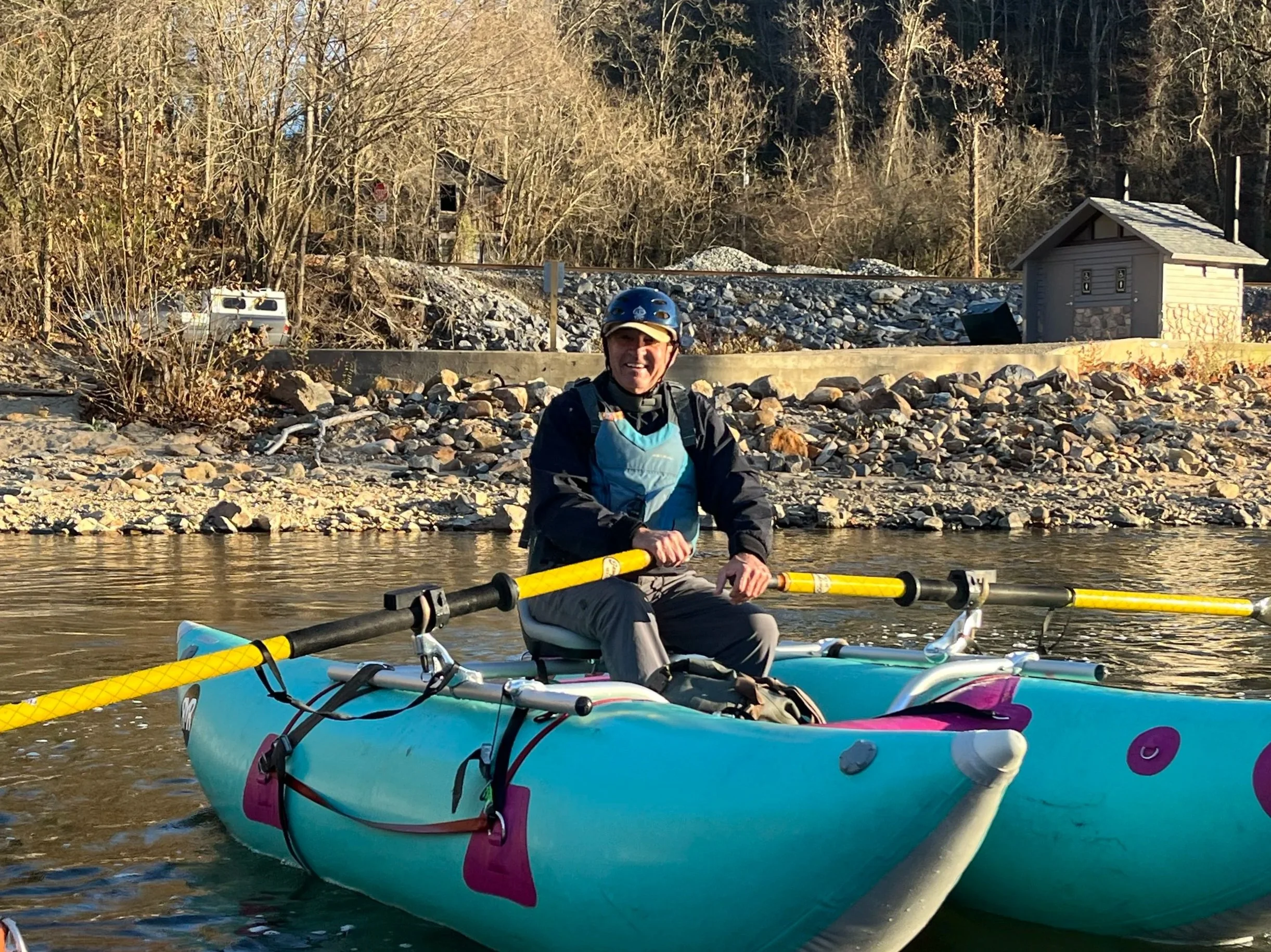 Person in a helmet and outdoor gear sitting in a blue and pink kayak on a river with trees and small buildings in the background.