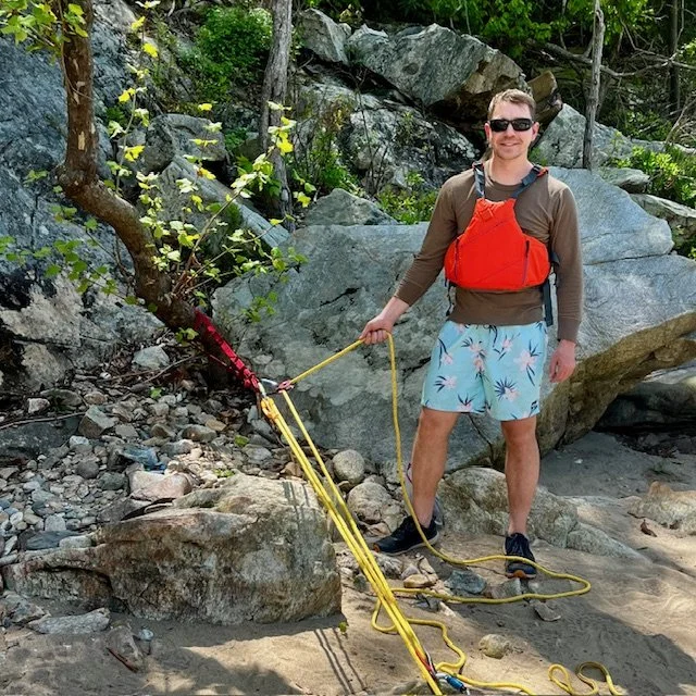 A man standing on a rocky beach holding a rope secured to a small tree, with an orange life jacket, sunglasses, brown long sleeve shirt, light blue shorts with a palm tree pattern, and sneakers, surrounded by rocks and greenery.