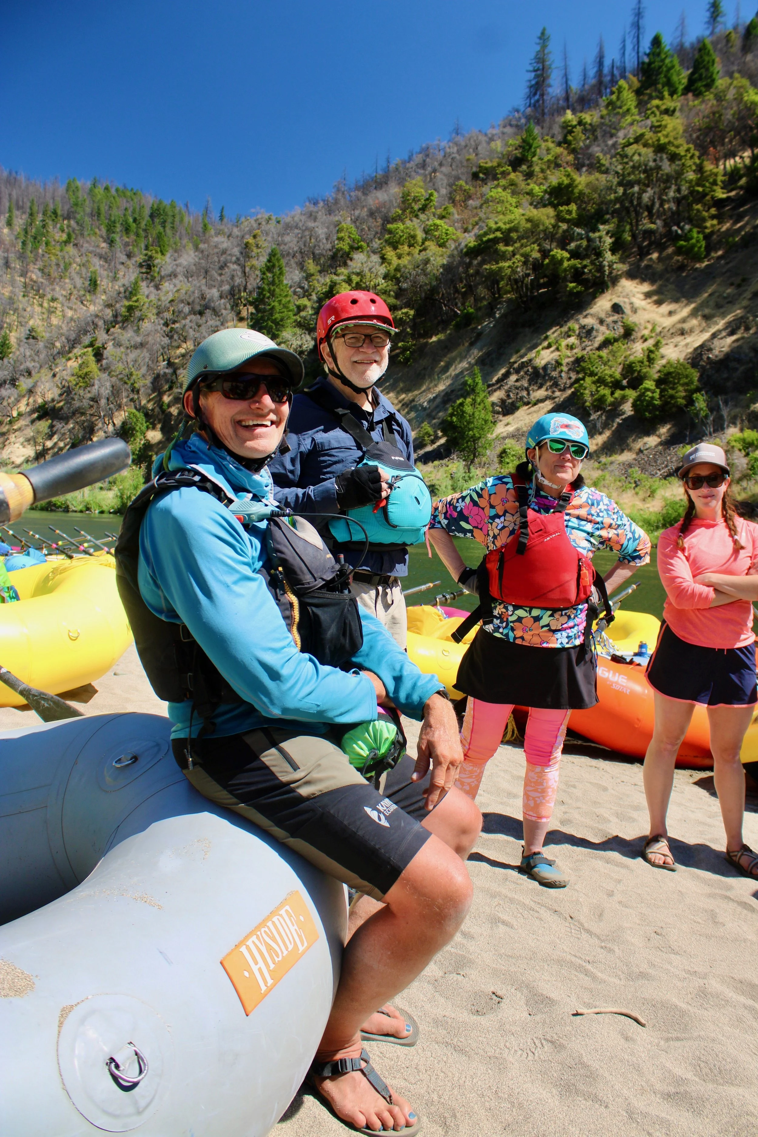 Group of four people wearing life jackets and helmets, standing on a sandy riverbank with inflatable kayaks nearby, surrounded by trees and hills under a clear blue sky.