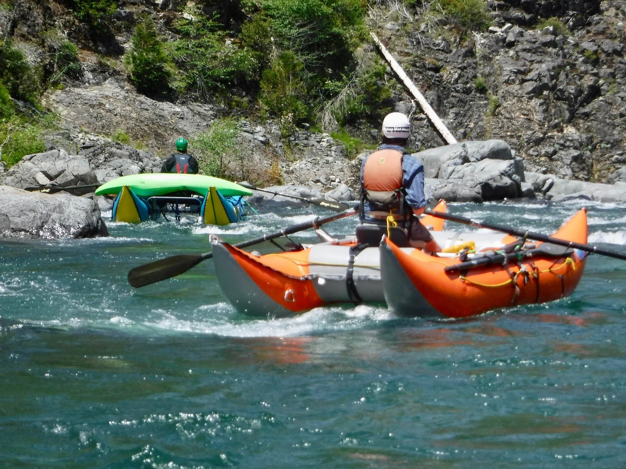 Two kayakers with helmets and life jackets navigating a rocky river with steep, wooded canyon walls, one kayak upside down.