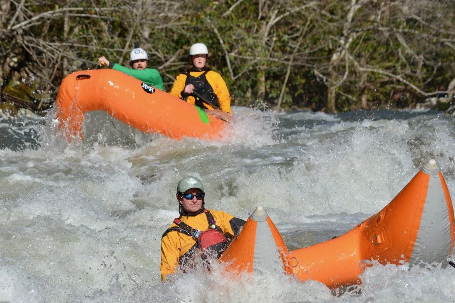 Three people white water rafting on a river, wearing helmets and life jackets, with two of them in an orange raft and one in the water near the raft.