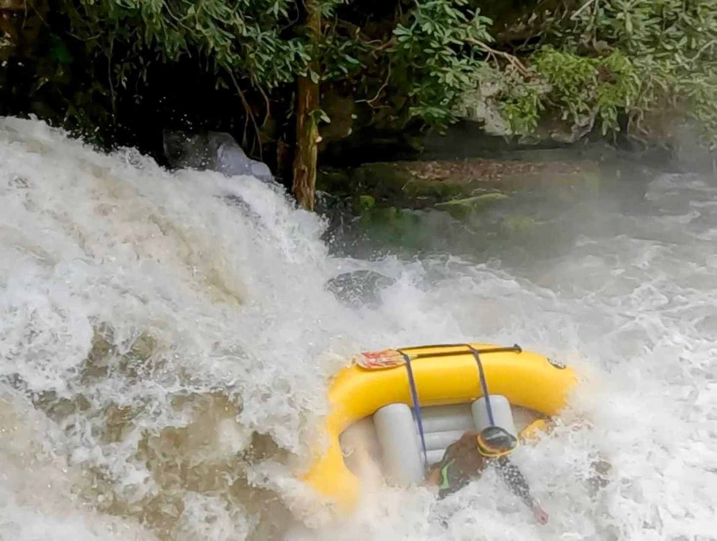 A person wearing a wetsuit and helmet trapped in a yellow inflatable raft amidst rushing whitewater surrounded by greenery.