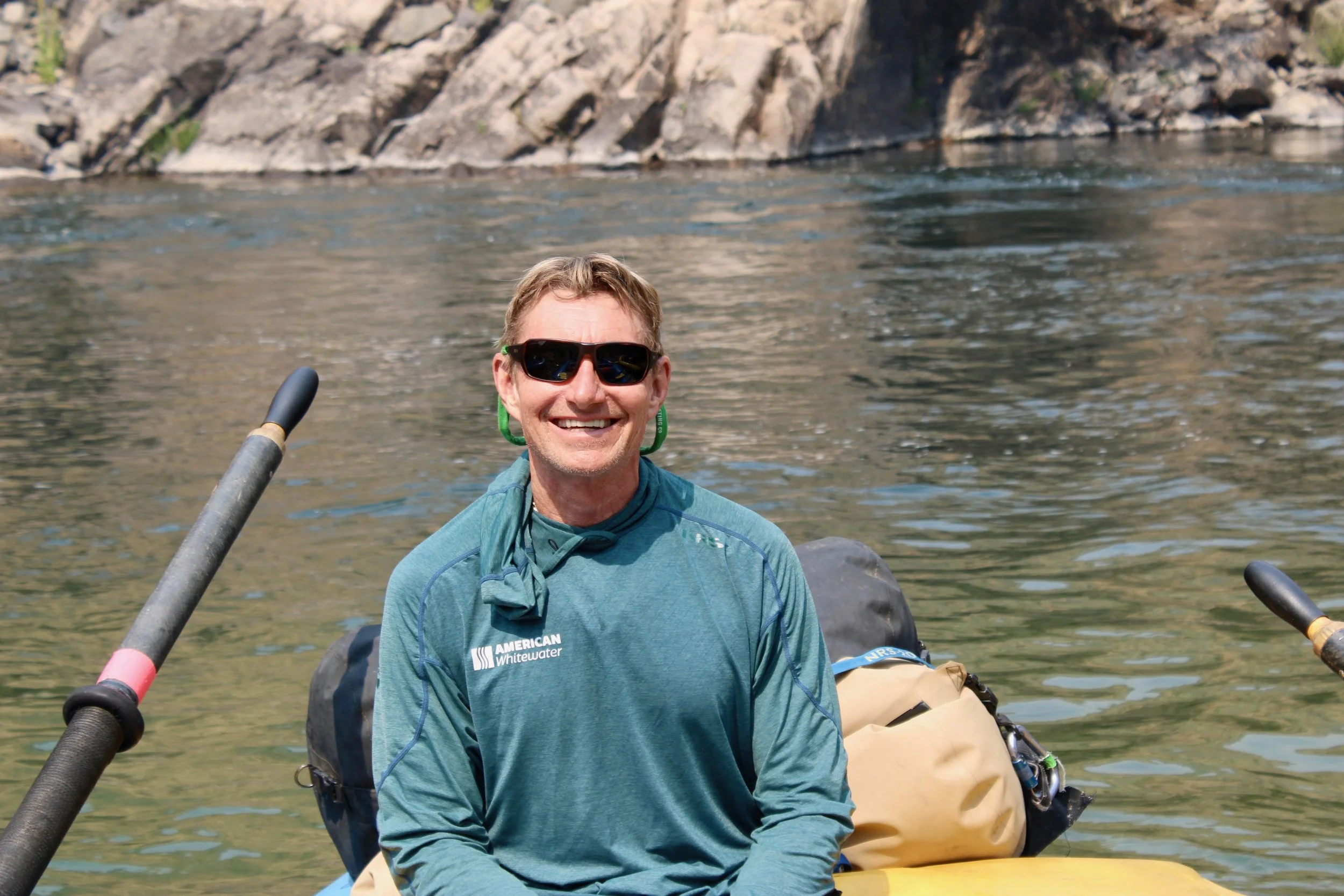 A smiling man wearing sunglasses kayaking on a river with rocky cliffs in the background.