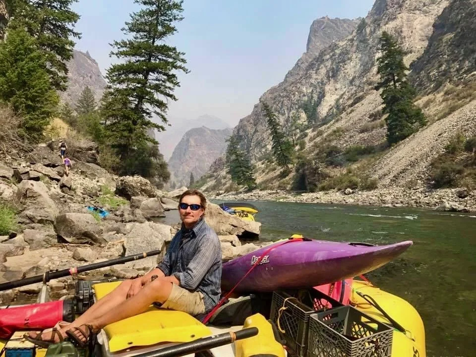 Man sitting on yellow inflatable kayak near river with mountains and trees in the background during daytime.