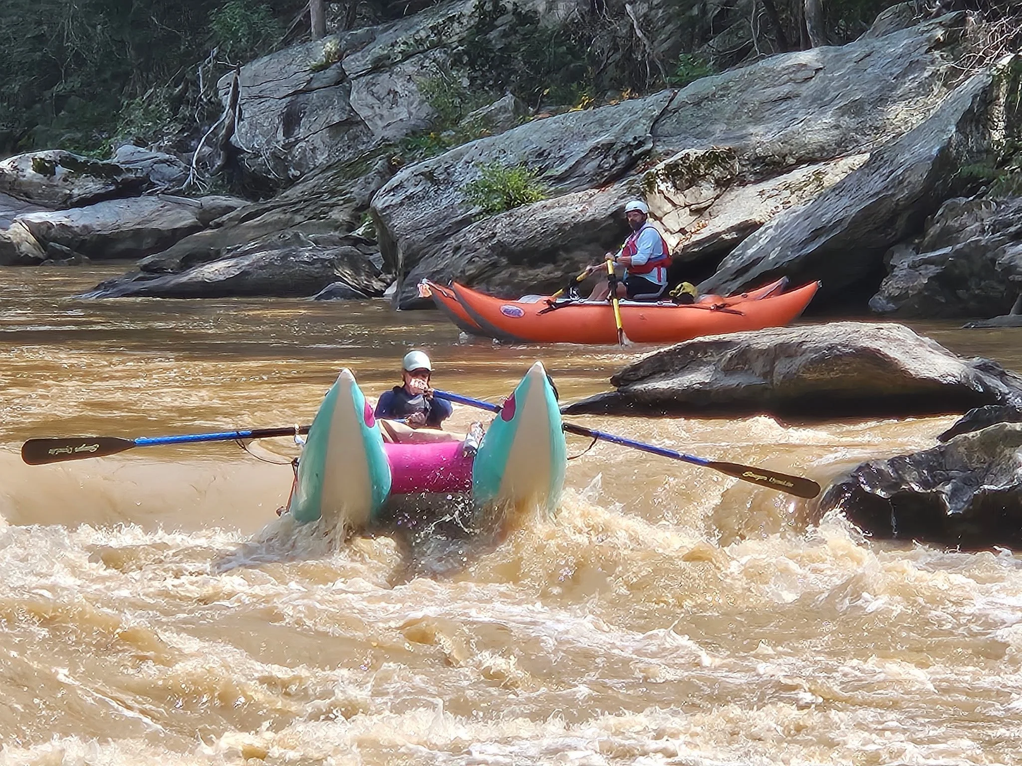 People kayaking on a river with large rocks and forested area in the background.