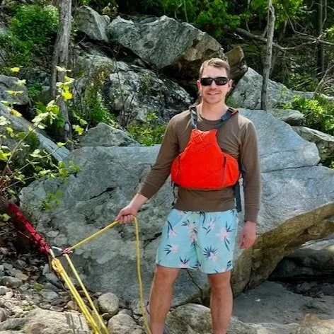 Man hiking outdoors with rocks and trees in the background, wearing sunglasses, a brown long-sleeve shirt, light blue shorts with a palm tree pattern, and carrying a bright orange backpack and yellow rope.