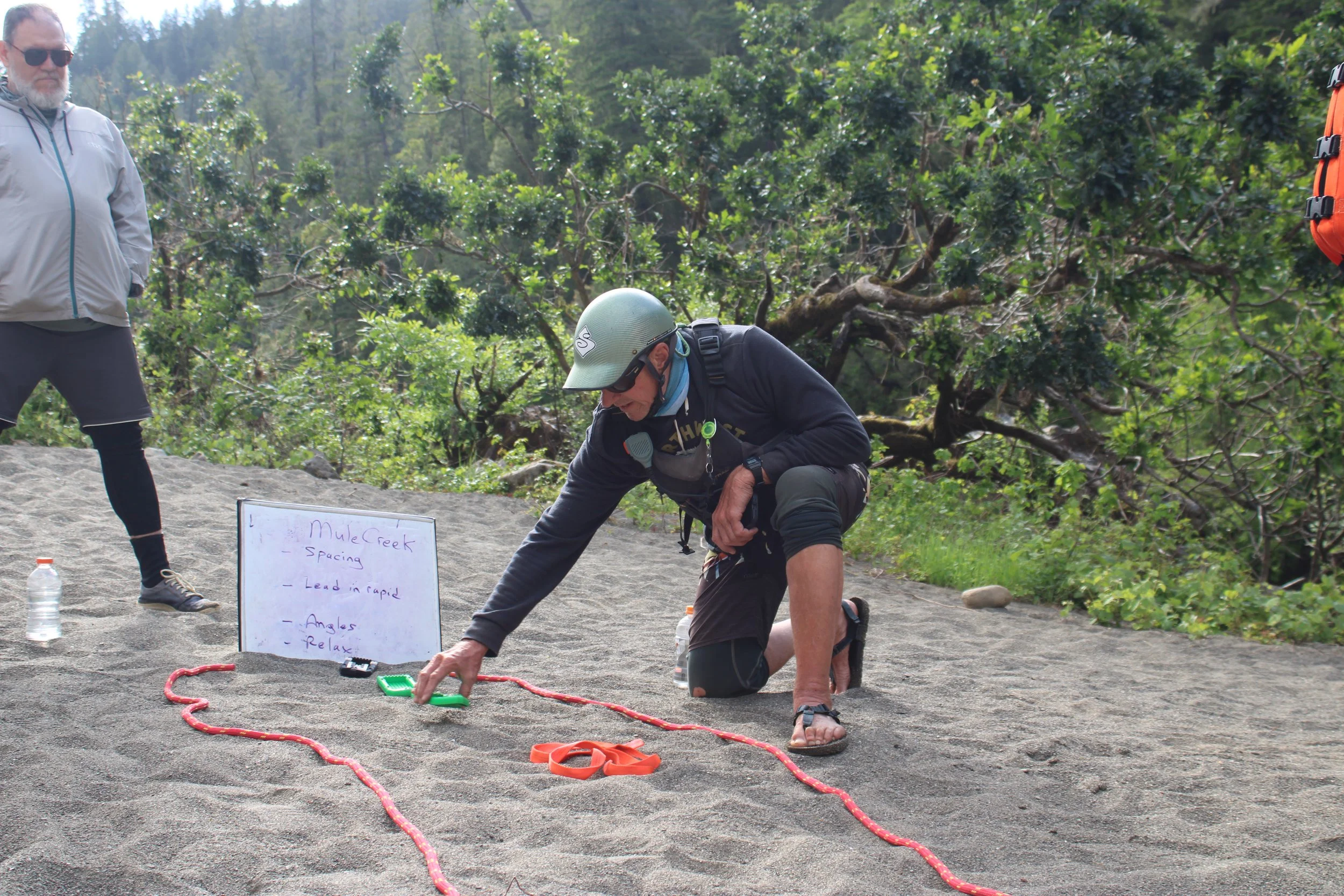 A man wearing a helmet and black clothing is kneeling on the ground, writing or placing an item inside a green container, with another man standing nearby in sunglasses and a gray jacket looking on. There are whiteboards, orange straps, and a water bottle on the ground, with a backdrop of lush green trees and a wooded area.