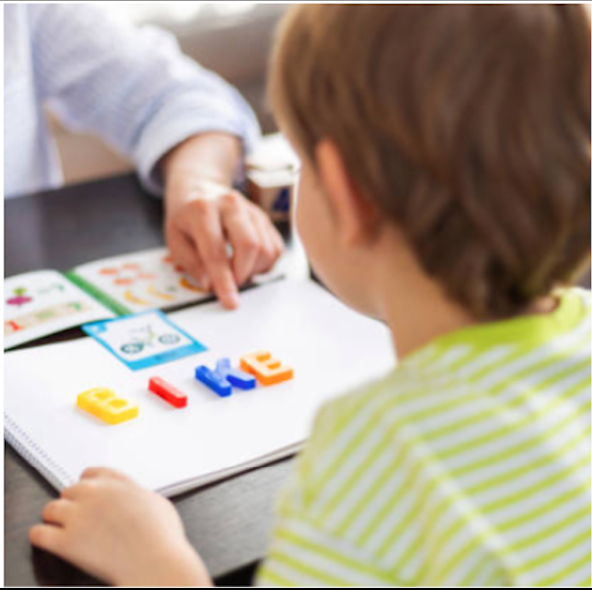 Child playing with colorful alphabet magnetic letters at a table with an adult pointing to a book.