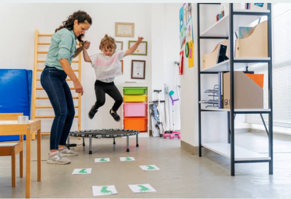 A woman and a girl jump on a small trampoline in a room with colorful storage bins and shelving, with activity signs on the floor.
