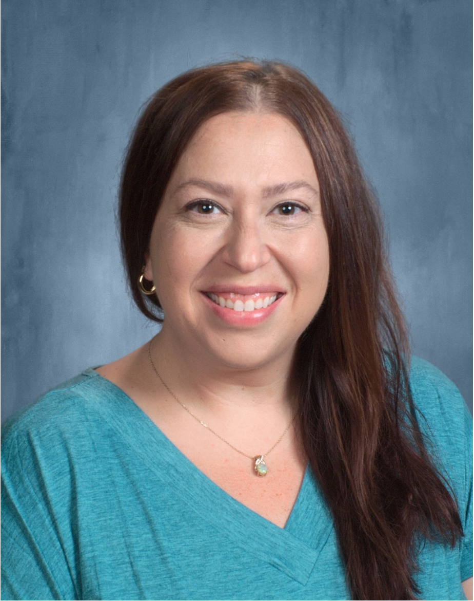A woman with long brown hair, wearing a blue top, earrings, and a necklace, smiling at the camera against a gray background.