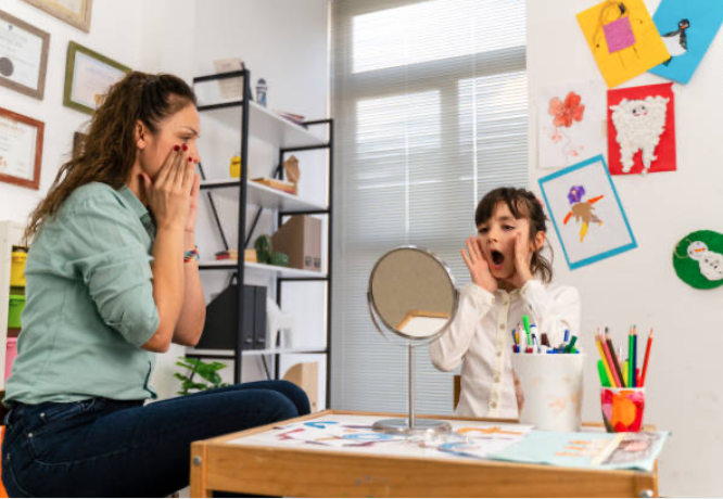 A young girl with brown hair, wearing a white shirt, sitting at a table with art supplies, reacting with surprise or excitement to her reflection in a small round mirror, in a colorful room with artwork on the walls.