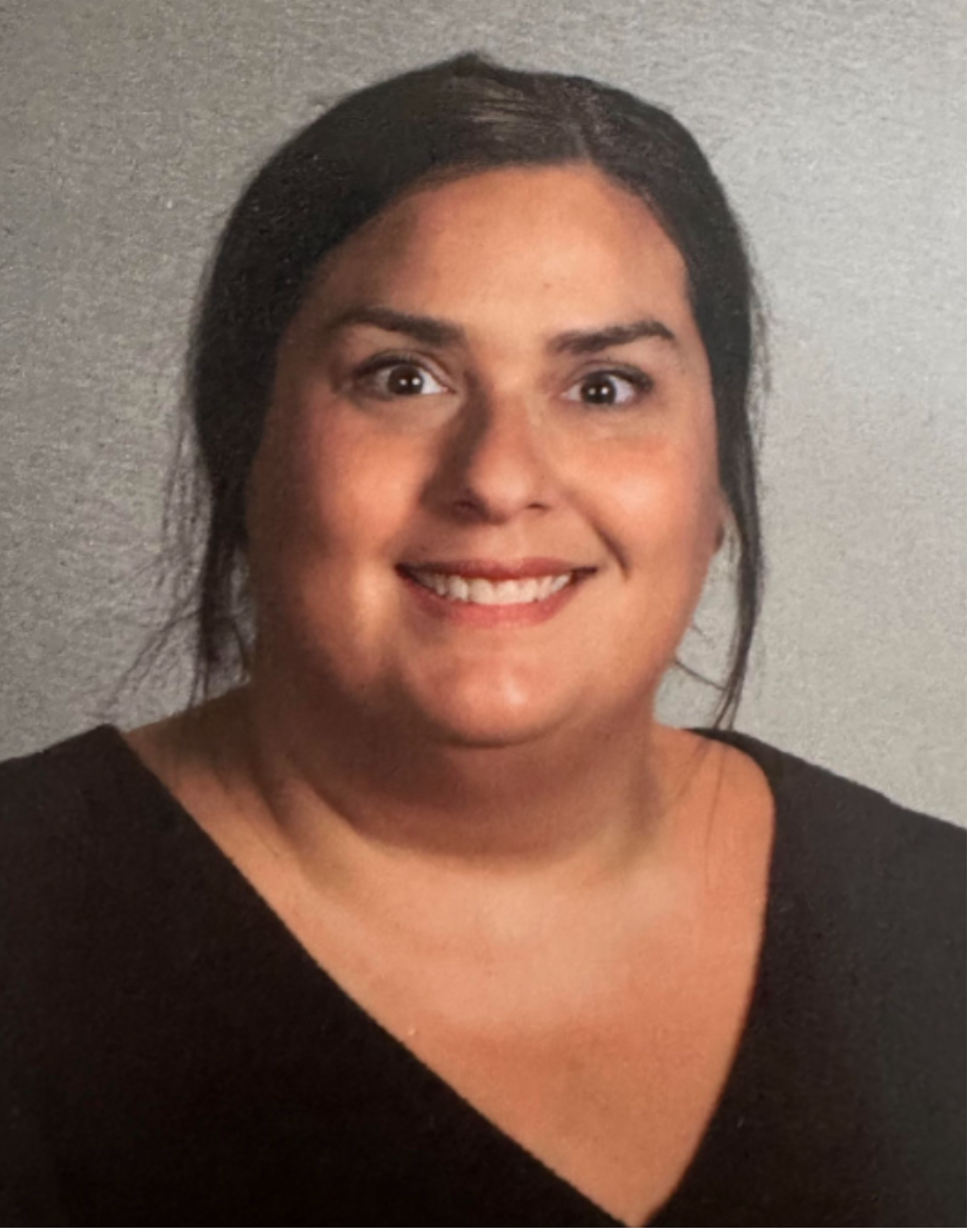 A woman with dark hair smiling at the camera, wearing a black top, standing against a plain wall background.