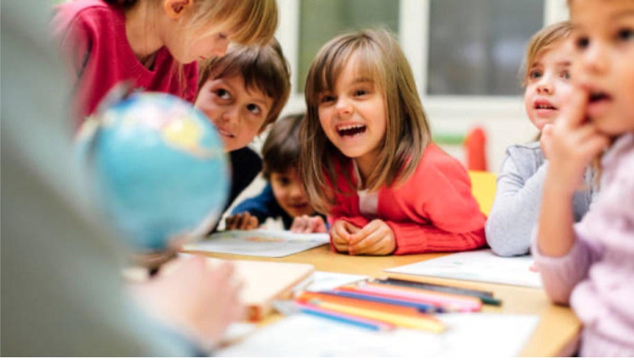 Young children and a teacher in a classroom looking at a globe on a table with colorful pens and papers.