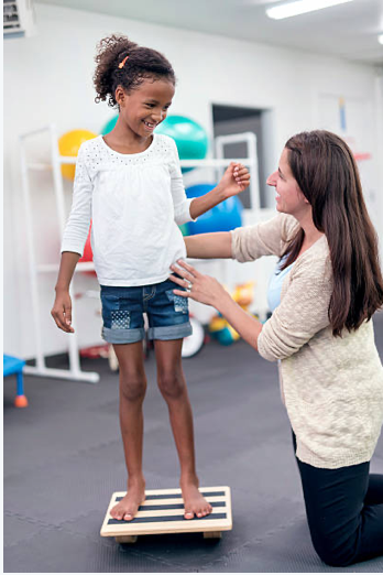Girl standing on a balance scale with a caregiver supporting her in a therapy or activity room.