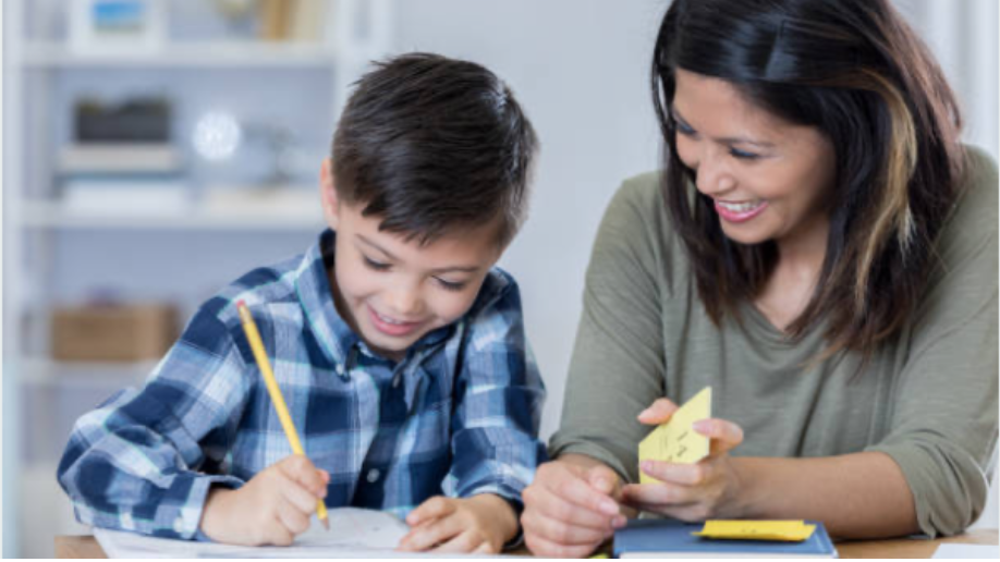 A boy and a woman sitting at a table, smiling and engaging in an activity. The boy is writing with a yellow pencil on paper, and the woman is holding a sticky note, sharing a moment of learning or playing together.
