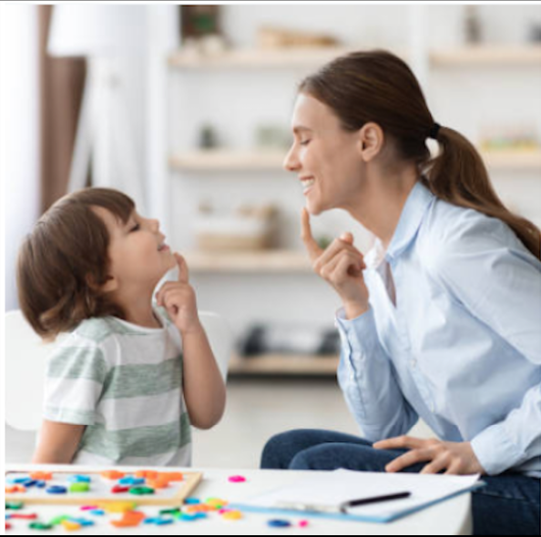 A woman and a young boy are sitting face to face, both smiling and touching their chins in a playful manner. They are indoors in a bright room with shelves in the background, and there are colorful game pieces and a notepad on the table between them.