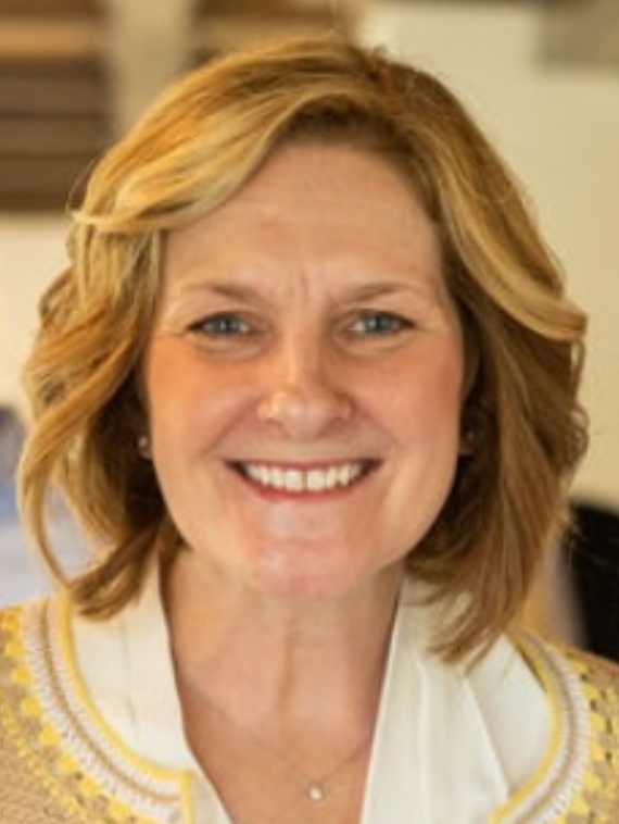 A smiling woman with blonde, wavy hair wearing a white top with yellow embroidery.