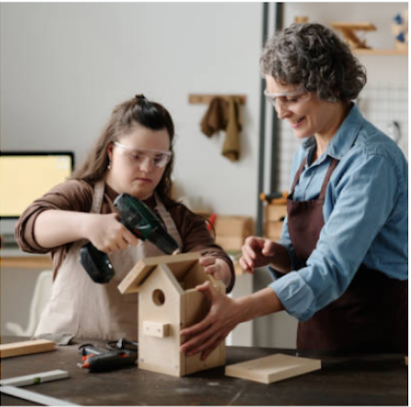 A young girl and an older woman, possibly a grandmother, working together on a woodworking project to build a birdhouse in a workshop. The girl uses a cordless drill while the woman guides her, both wearing safety glasses.