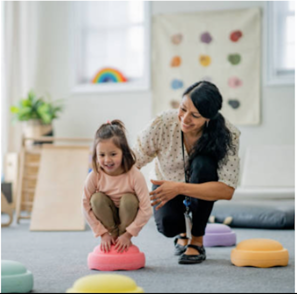 A young girl sitting cross-legged on a pink cushion with a woman, in a bright therapy or activity room. The girl is smiling, and the woman is engaging with her, with colorful cushions and a wall chart with colorful shapes in the background.