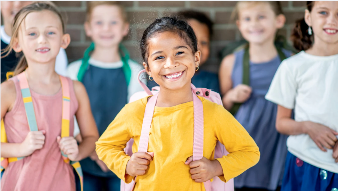 A group of diverse children smiling and standing outside, wearing backpacks.