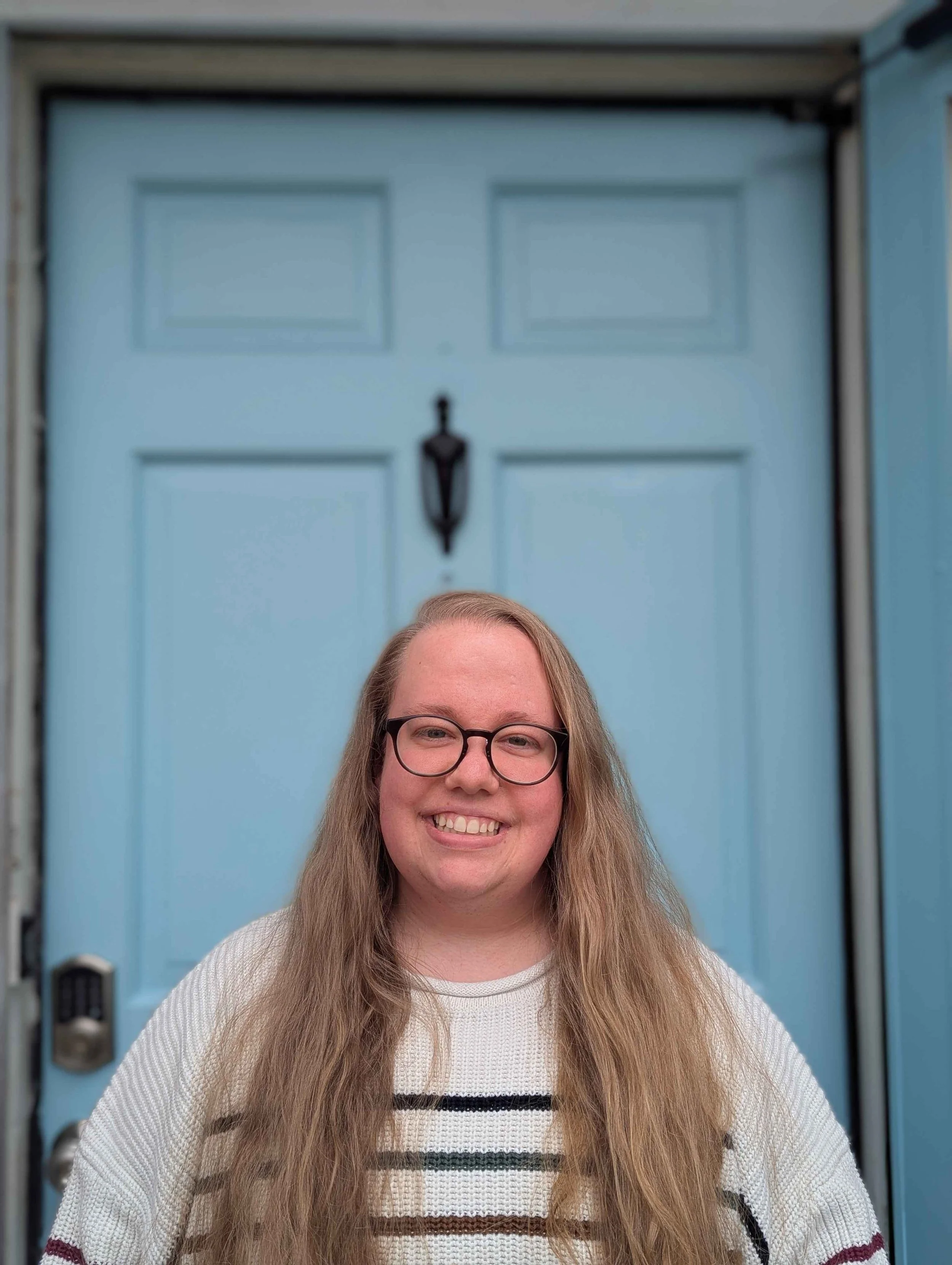 A smiling woman with long long wavy hair and glasses standing in front of a blue door with a black handle and mail slot.