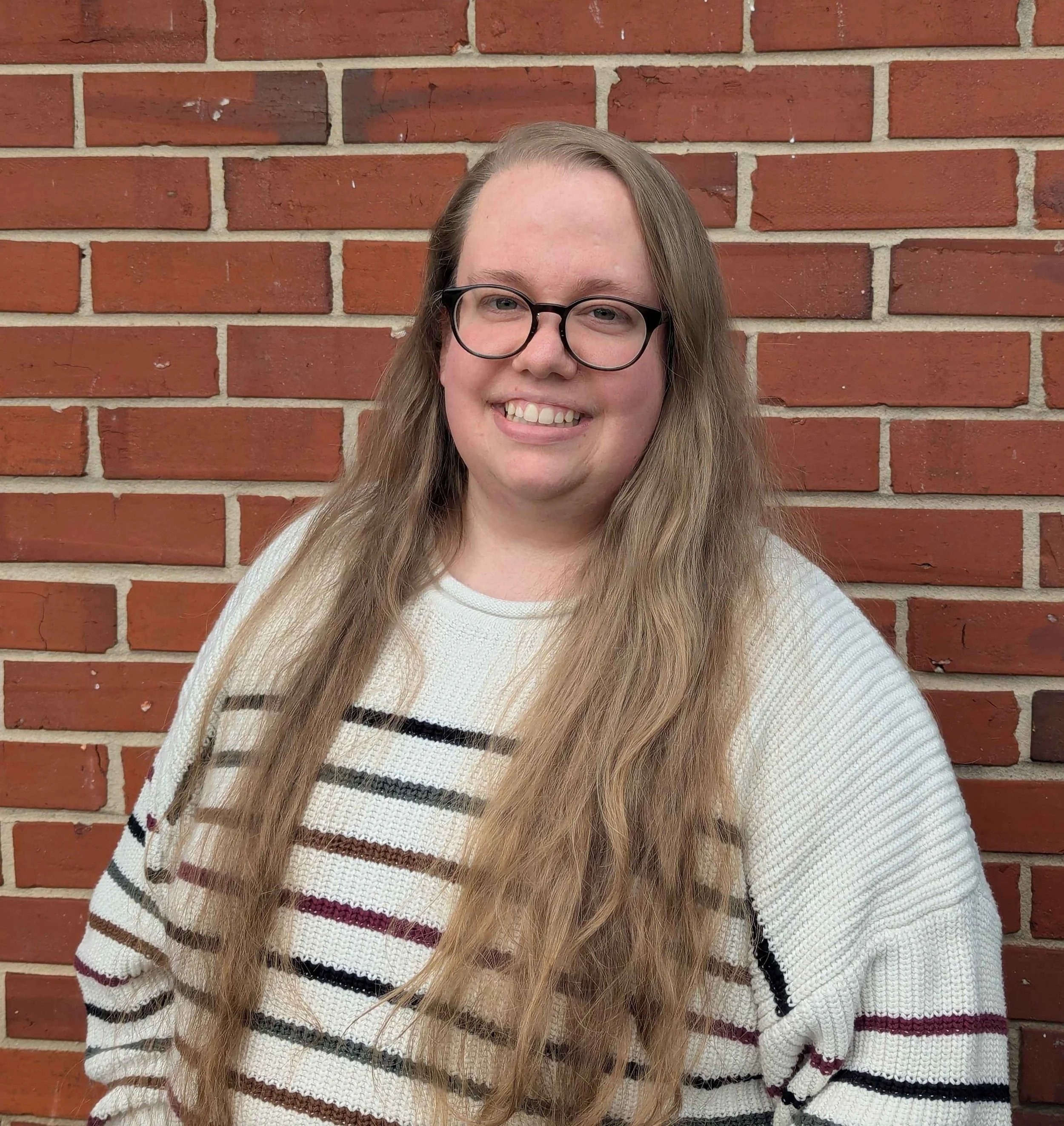 A young woman with long wavy hair wearing glasses and a striped sweater, standing in front of a brick wall and smiling.