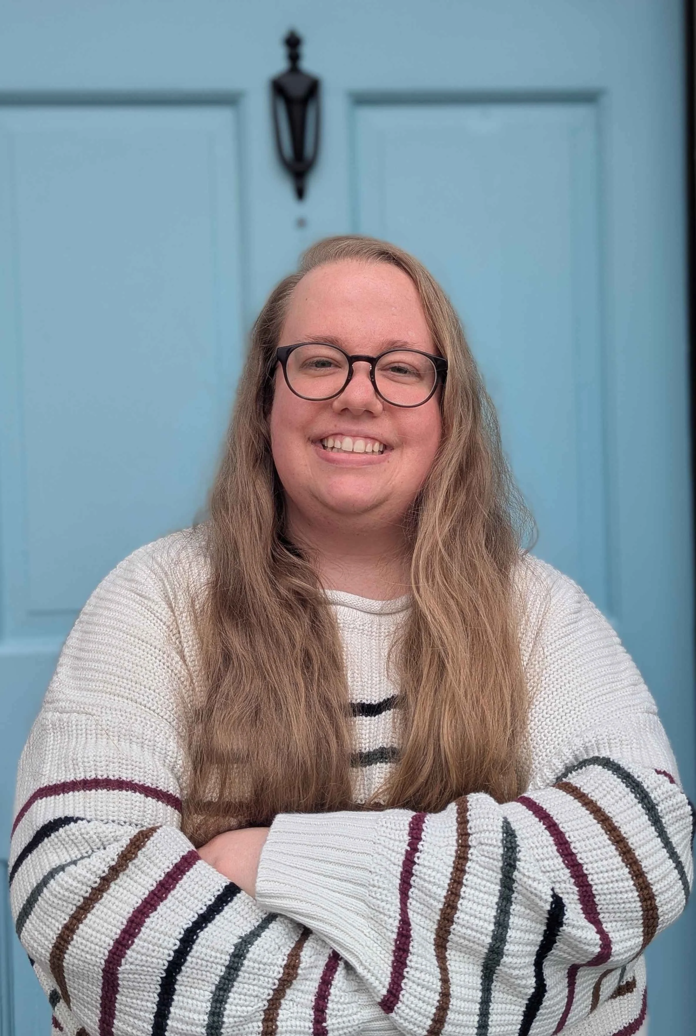 A smiling woman with long wavy hair and glasses, wearing a white sweater with multicolored stripes, standing in front of a light blue door with a black lantern.