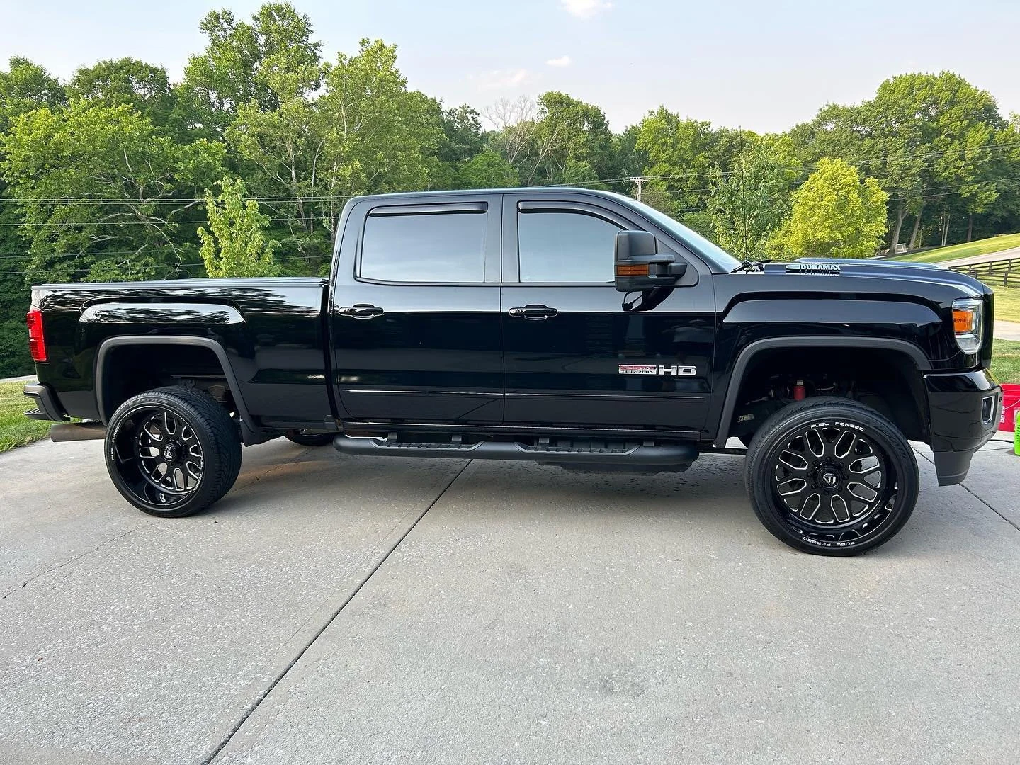 Black GMC Sierra HD pickup truck parked on a concrete driveway with grass and trees in the background.