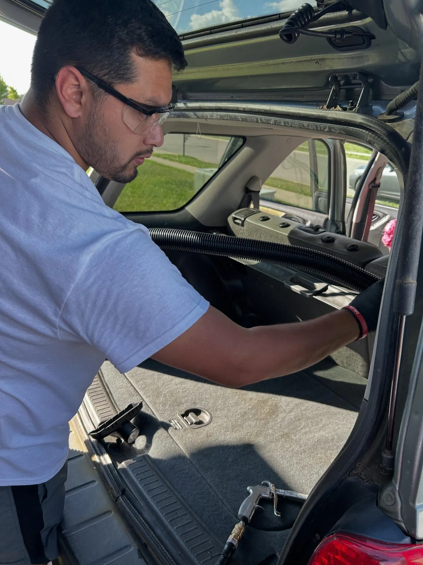 A man wearing safety glasses and gloves working inside the trunk of a vehicle, possibly repairing or inspecting the vehicle.