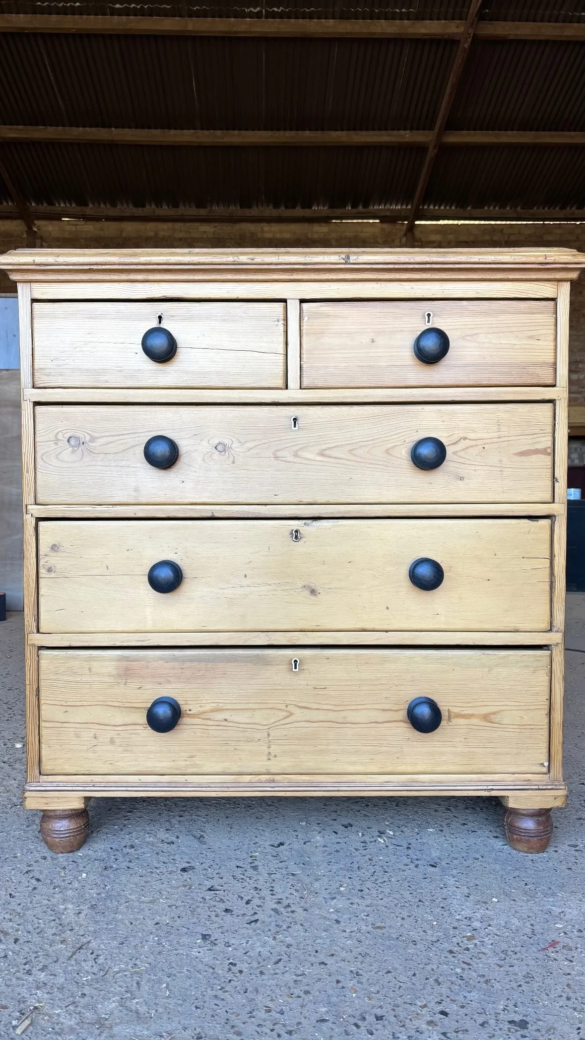 A 19th century Victorian pine chest of drawers with five large drawers, each with a black round knob, set on a rough concrete surface in a warehouse or barn with a metal roof overhead. Bring It Home Furniture, Hingham Norfolk.