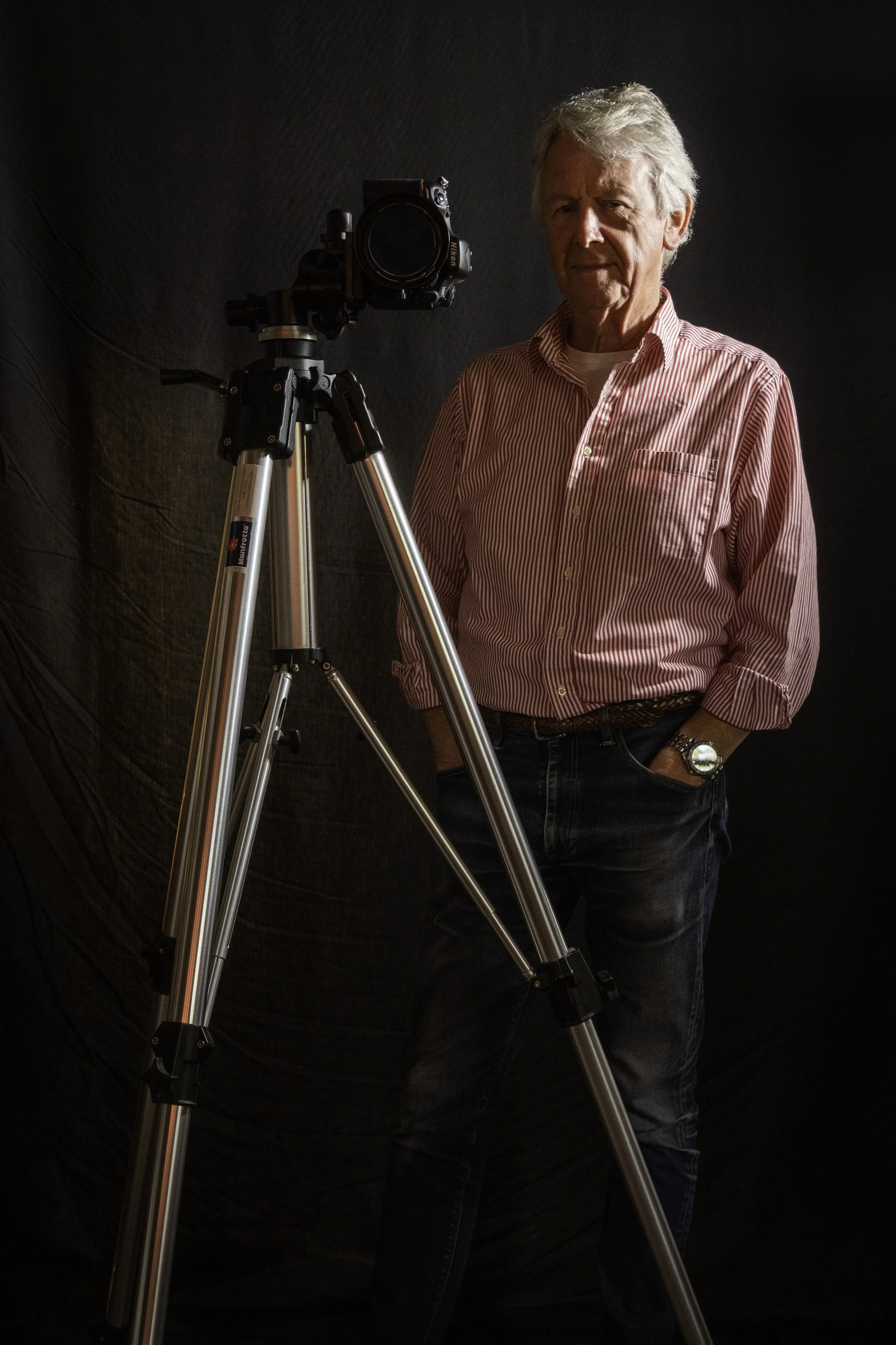 The photographer stands next to a camera mounted on a tripod in front of a dark background.
