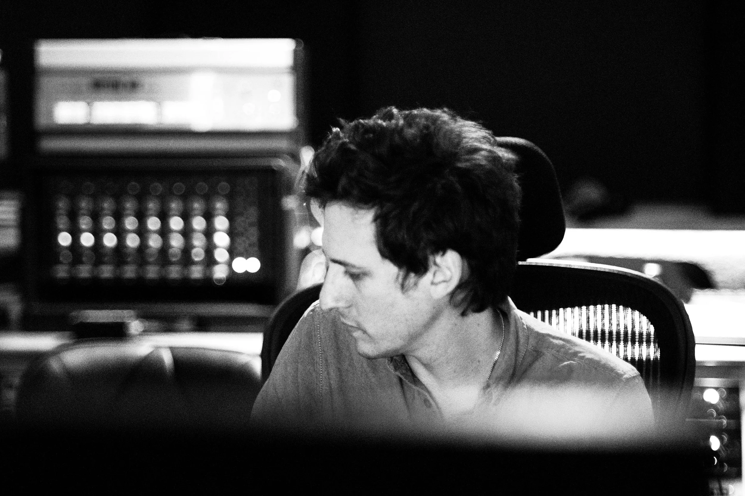 Black and white photo of a young man with curly dark hair sitting in a chair, focused on something in front of him, with audio equipment visible in the background.