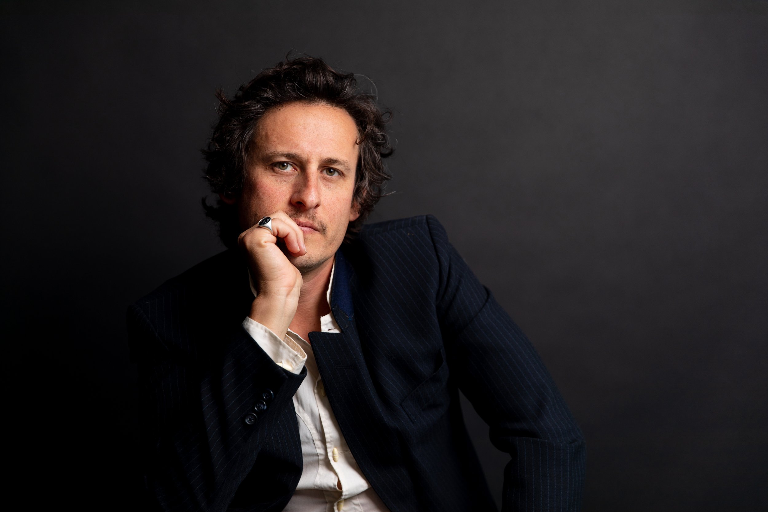 Scott Hirsch in a dark blazer and white shirt, sitting against a dark background, with his hand resting on his face, looking into the camera.