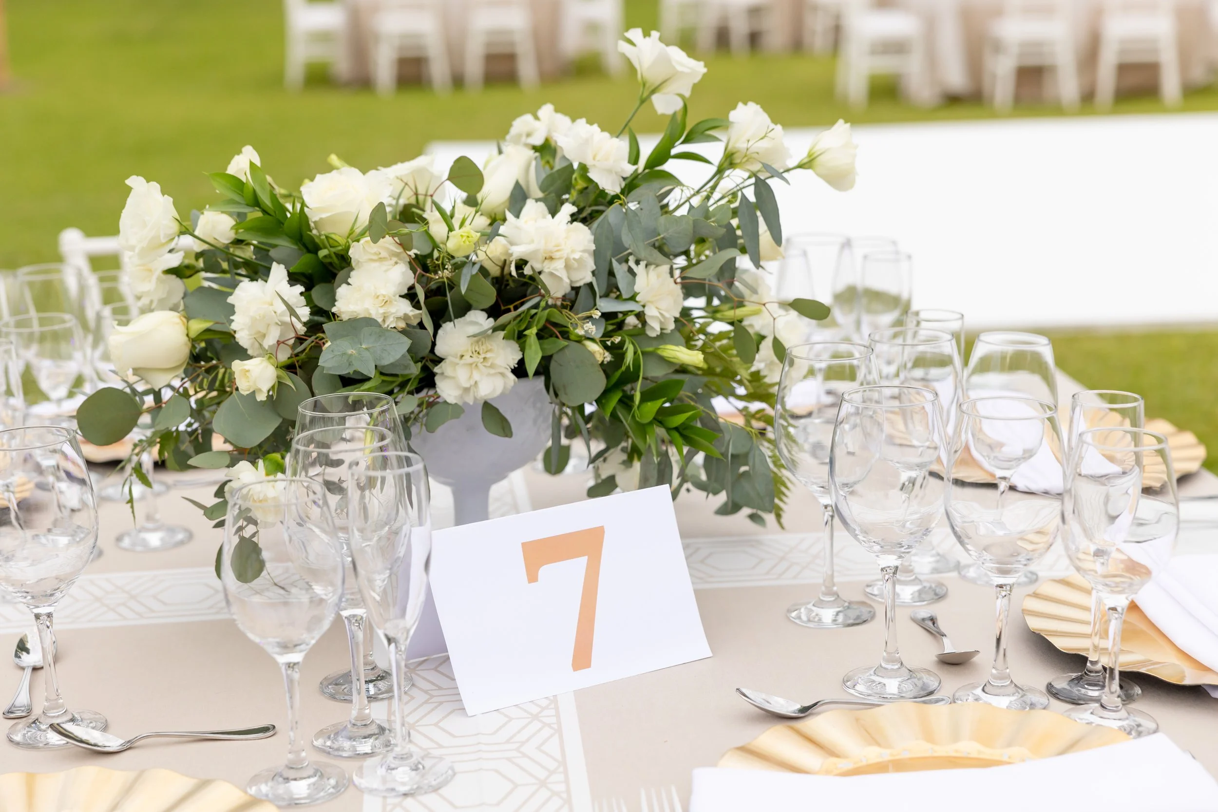 Centre de table avec fleurs blanches et verdure, assiettes dorées, verres à vin, et éléments décoratifs, installation pour un événement extérieur.