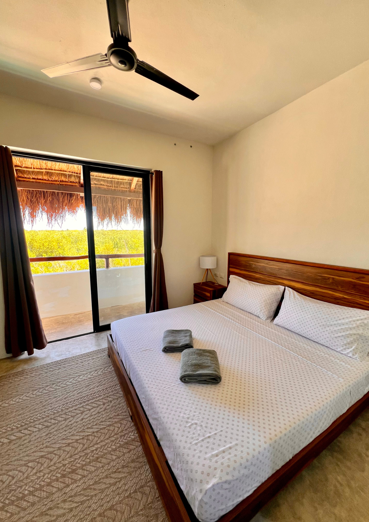 Chambre d'hôtel avec un lit double, deux serviettes pliées dessus, une table de chevet avec une lampe, un balcon avec des portes en verre, vue sur la végétation, et un plafond avec un ventilateur.