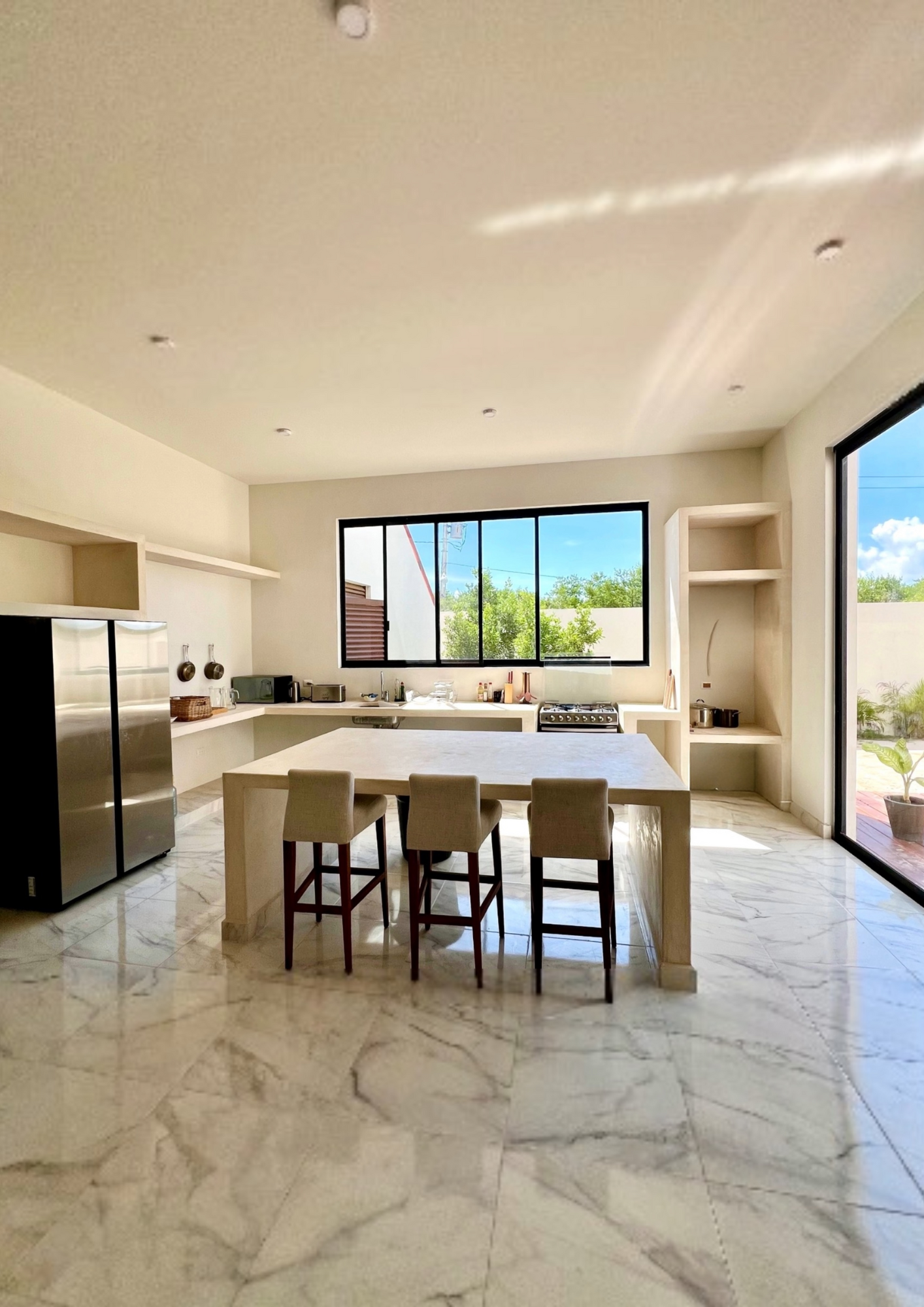 Cuisine moderne lumineuse avec une grande table centrale, quatre chaises, un réfrigérateur en acier inoxydable, une fenêtre large laissant entrer la lumière naturelle, étagères encastrées, sol en marbre clair, et un espace extérieur visible par une p