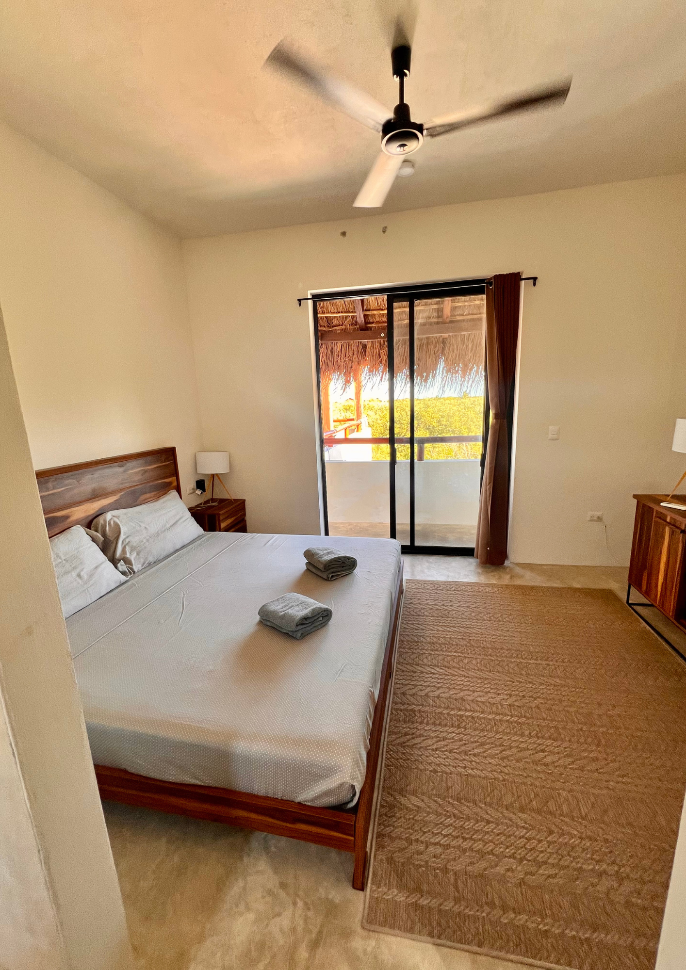 Chambre avec lit double, deux serviettes pliées dessus, une table de nuit avec lampe, porte-fenêtre menant à une terrasse avec vue sur la nature, ventilateur au plafond.