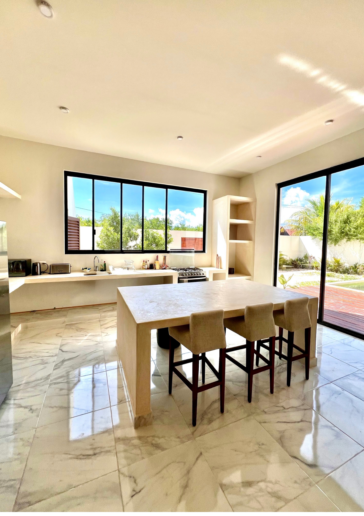 Cuisine moderne avec grande table en marbre, quatre chaises en tissu beige, fenêtres en verre laissant entrer la lumière naturelle, vue sur jardin extérieur verdoyant.