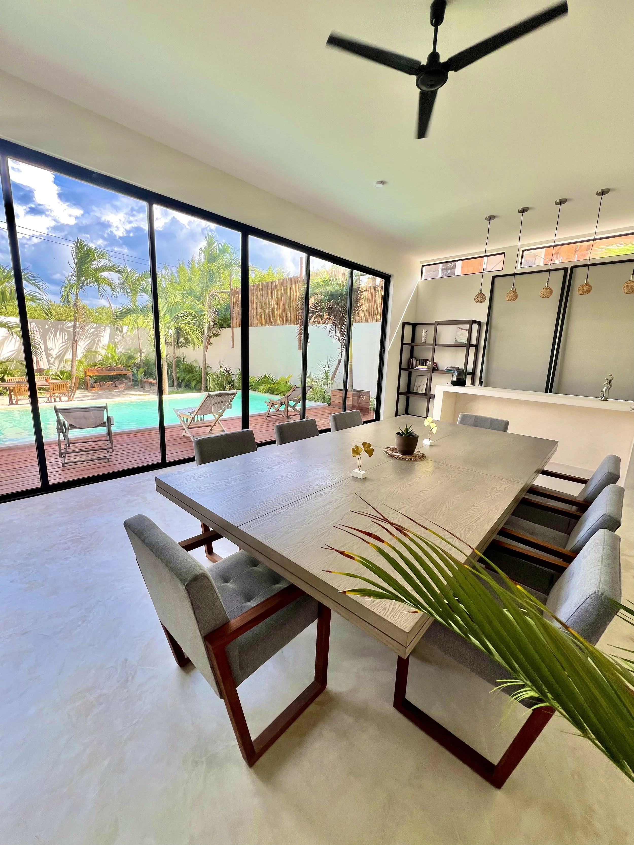 Salle à manger moderne avec grande table en bois, huit chaises en tissu gris, décoration minimaliste, jardin avec piscine visible par des grandes fenêtres en arrière-plan