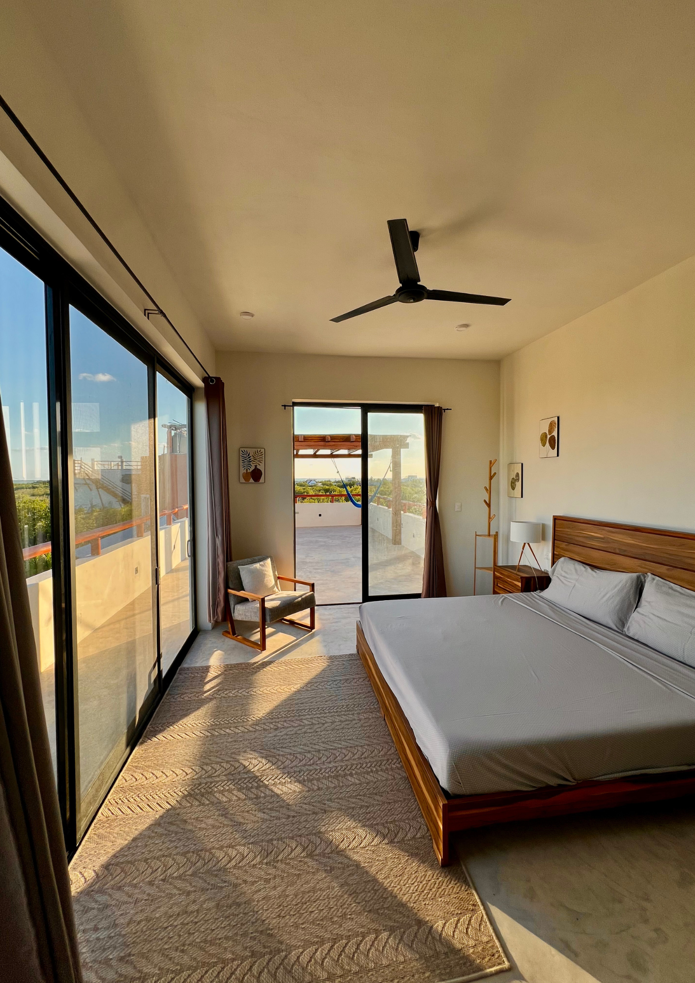Chambre lumineuse avec un lit en bois, grandes fenêtres donnant sur un balcon extérieur, un fauteuil, un lampadaire, décorations murales, et un plafond avec une grille ventilateur.
