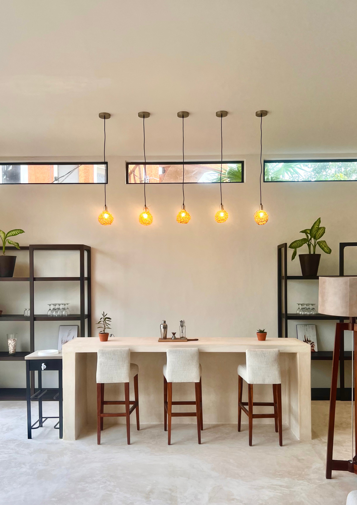Une salle à manger moderne avec un grand comptoir blanc, trois chaises en bois avec coussins clairs, et des étagères noires avec des plantes et des accessoires. Quatre pendentifs suspendus au plafond diffuses une lumière chaude. Fenêtres horizontales