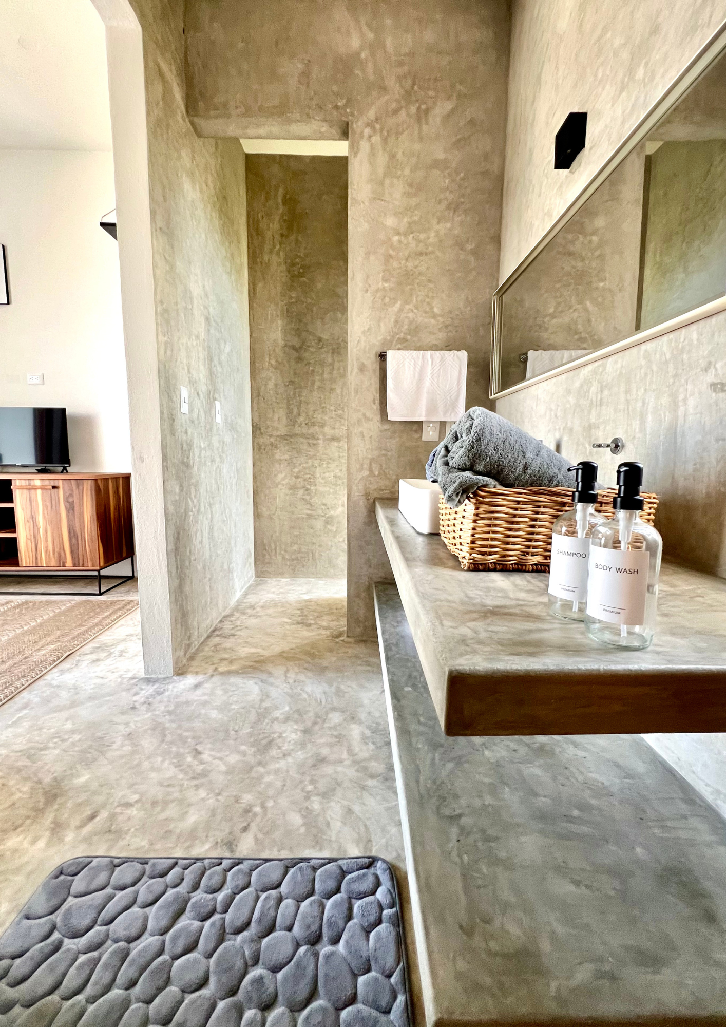 Intérieur moderne avec un mur en béton, une table avec des serviettes, des bouteilles de shampoing et de gel douche, un grand miroir, et une télévision en bois dans le fond.