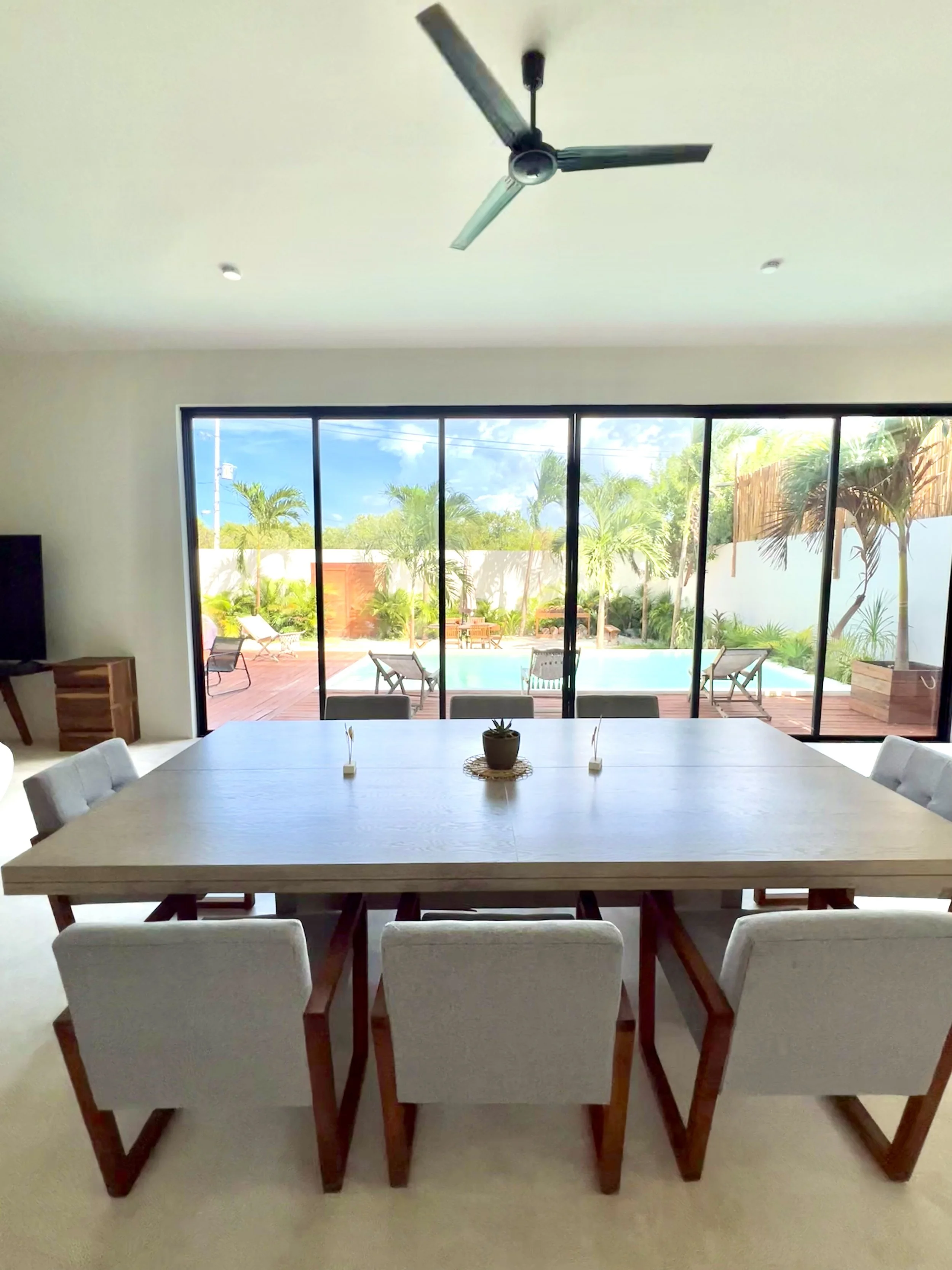 Salle à manger moderne avec table en bois clair, six chaises rembourrées, vue d'une piscine et d'un jardin tropical par de grandes fenêtres en verre.