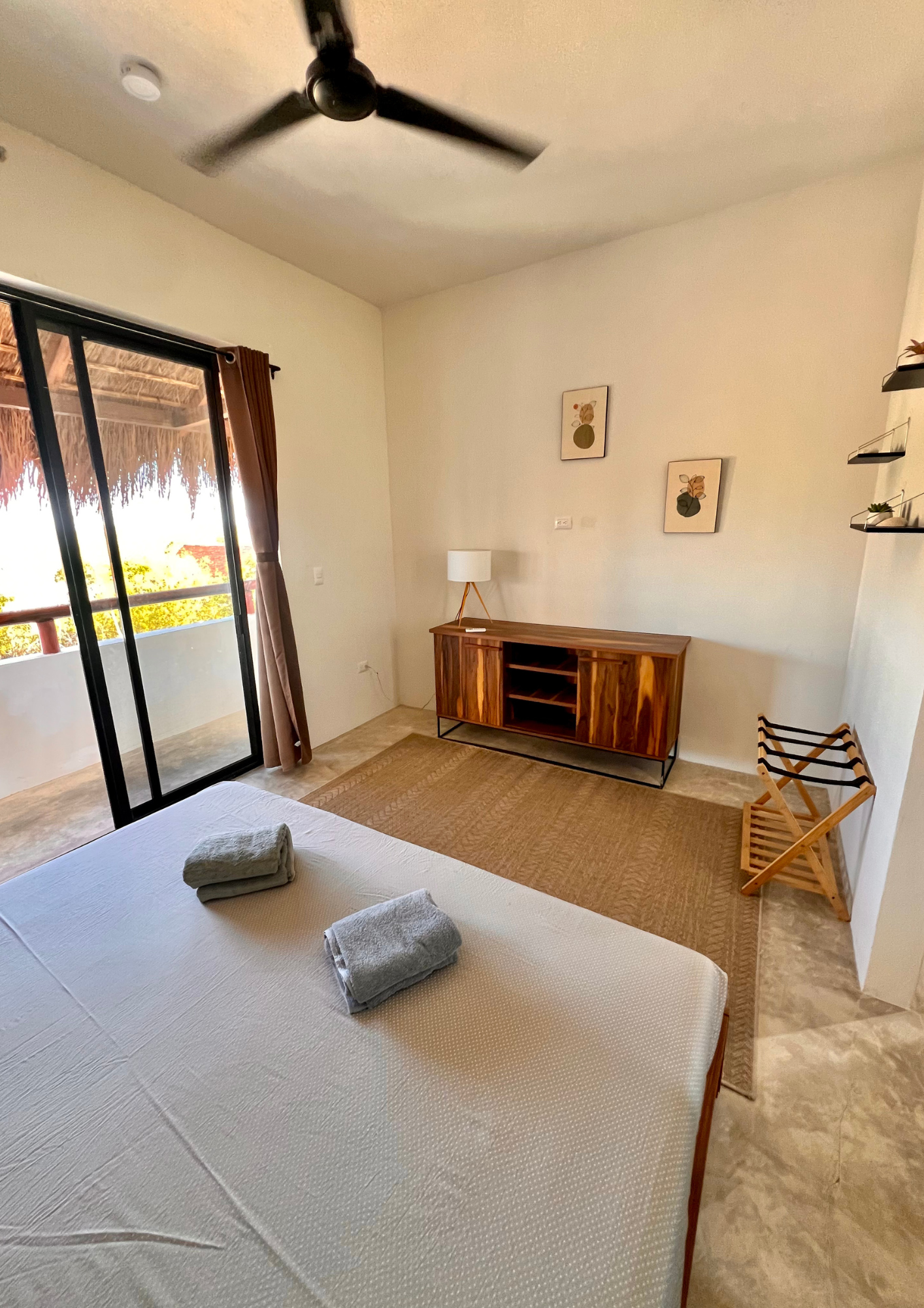 Chambre avec un lit fait, deux serviettes pliées dessus, une grande fenêtre avec une porte-fenêtre donnant sur un balcon, un meuble en bois avec une lampe à côté, deux tableaux avec des illustrations florales, un tapis en jute, une assise en bois pou